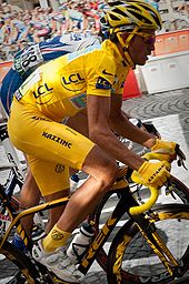 A man in yellow clothes, with a yellow helmet and yellow handgloves, riding a bicycle. Just behind him is another cyclist, in blue clothes.