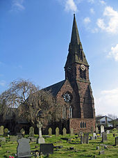 The west end, with a rose window, and the steeple of a Gothic style church, with a clock face; gravestones in the foreground and a leafless tree to the left.
