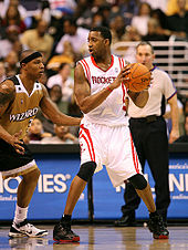 A basketball player, wearing a white jersey with the word "ROCKETS" across the front, holds a basketball away from another basketball player guarding him. A referee stands in the background.