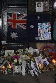 Dark doorway and doorstep with section of footpath. On the lower part of the dark door is a partly obscured Australian flag with dark blue background; red and white crosses on top left, large white star underneath and three white stars at the right with others obscured. Obsuring the right side of the flag is a white sheet with light blue writing, "Love + Miss You Always Heath" with two red hearts nearby, other writing includes "I'll never quit you" in darker blue, more writing is indistinct. This sheet also has six photos of a man. Above the flag, on the door is a smaller sheet with a photo of a man and indistinct writing below. On the doorstep and section of footpath are some 14 groups of flowers wrapped in plastic or paper, together with nine or so cards, five or six lit candles, and more photos. On the right side is a straw hat.