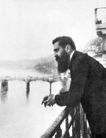 A long-bearded man in his early forties leaning over a railing with a bridge in the background.
