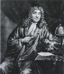 A man wearing a long, curly wig and a full robe is sitting, looking out. His left arm rests on a small table, with his left hand holding a box. Behind him is a globe