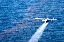 A large four propeller airplane spraying liquid over oil-sheen water