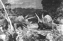 A black-and-white screenshot of three triceratops atop a cliff with trees surrounding them and mountains in the background.
