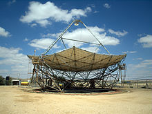 A horizontal parabolic dish, with a triangular structure on its top. Around it is a flat sandy area, with desert in the background. It's a sunny day, with a few white clouds in the blue skies.