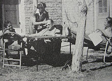 Three men sit in chairs outside a house, a bald man in his forties is sitting in the centre next to a table between the two younger men. The man on the right has his feet on the table and is holding a screenplay. the older man is having his brow wiped by a nurse who is standing over him.
