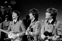 A black-and-white image of three men playing guitar. They are wearing grey buttoned-up suit jackets with ties underneath. An audience is visible behind them on the left.